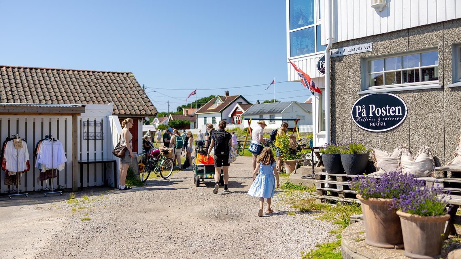 Folk og barn rusler mellom små butikker og kaféen "På Posten" på Herføl en solfylt dag.