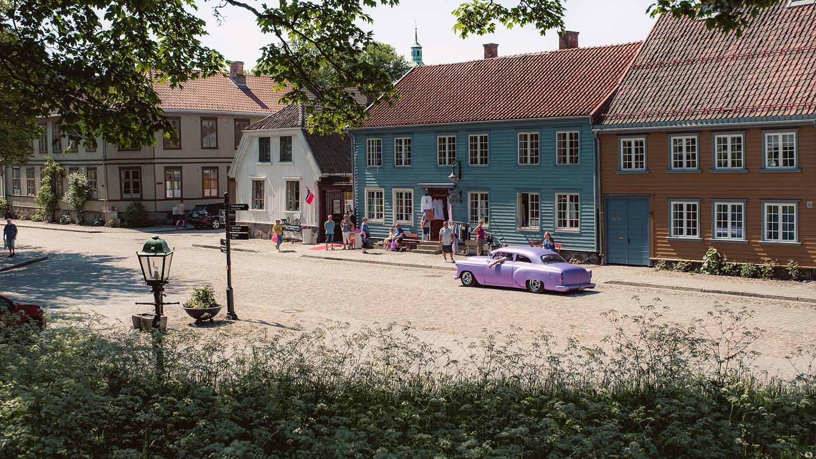 Brosteinsgate i Gamlebyen i Fredrikstad med fargerike trehus, folk som rusler og en lilla veteranbil parkert i solen.