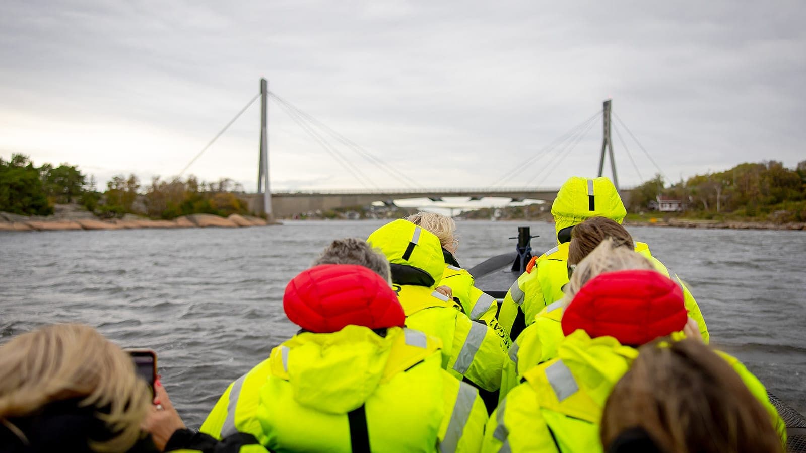 Gruppe i gule flytedrakter på RIB-tur i Hvalerskjærgården en høstdag, med Puttesund bro i bakgrunnen.