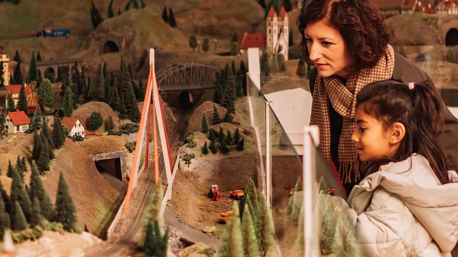 En voksen og et barn ser på et detaljert modelljernbaneanlegg med landskap, broer og små hus.