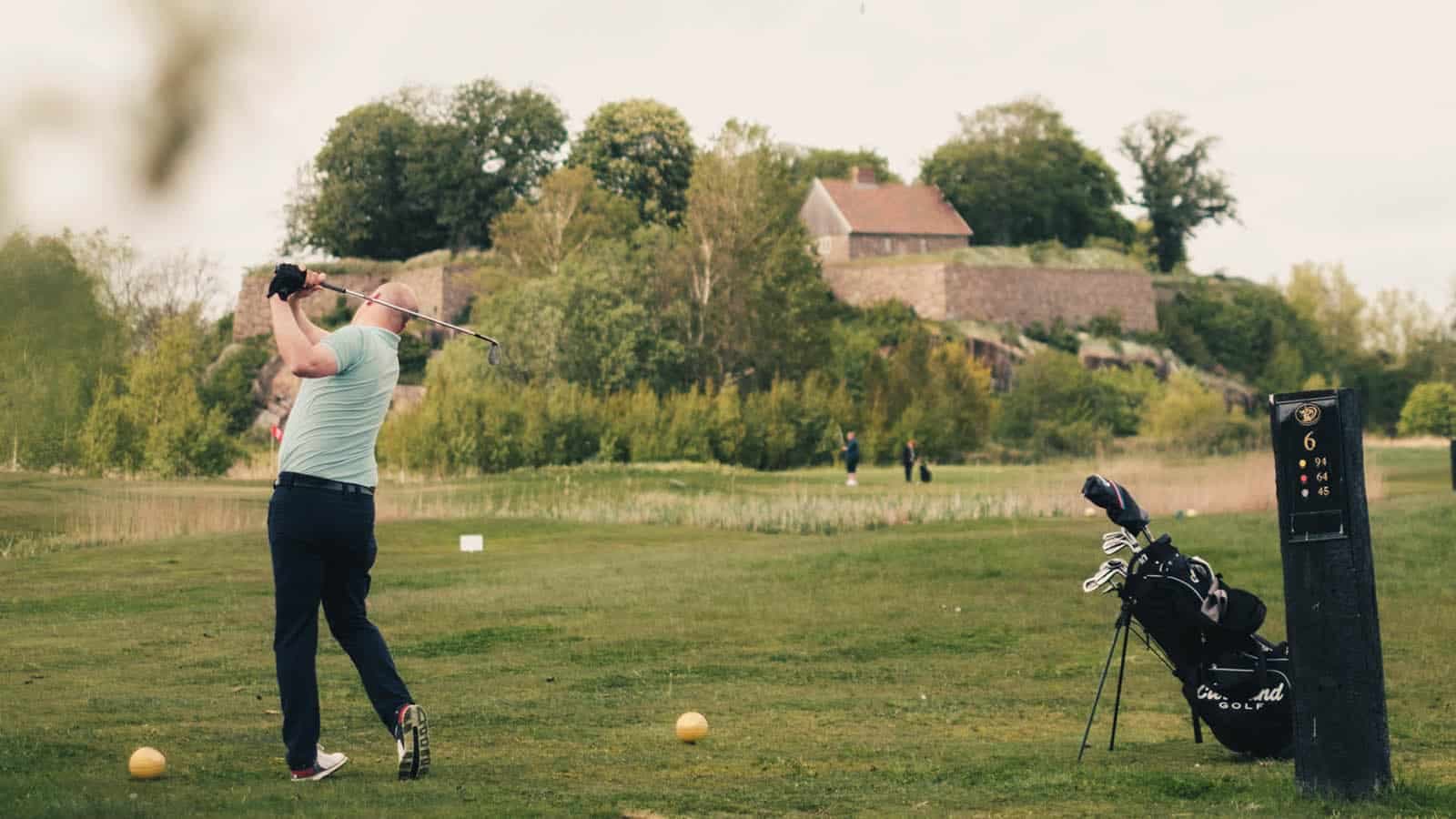 Golfspiller slår ut på banen ved Kongsten fort, Gamle Fredrikstad Golfklubb, med golfbag og historiske murer i bakgrunnen.