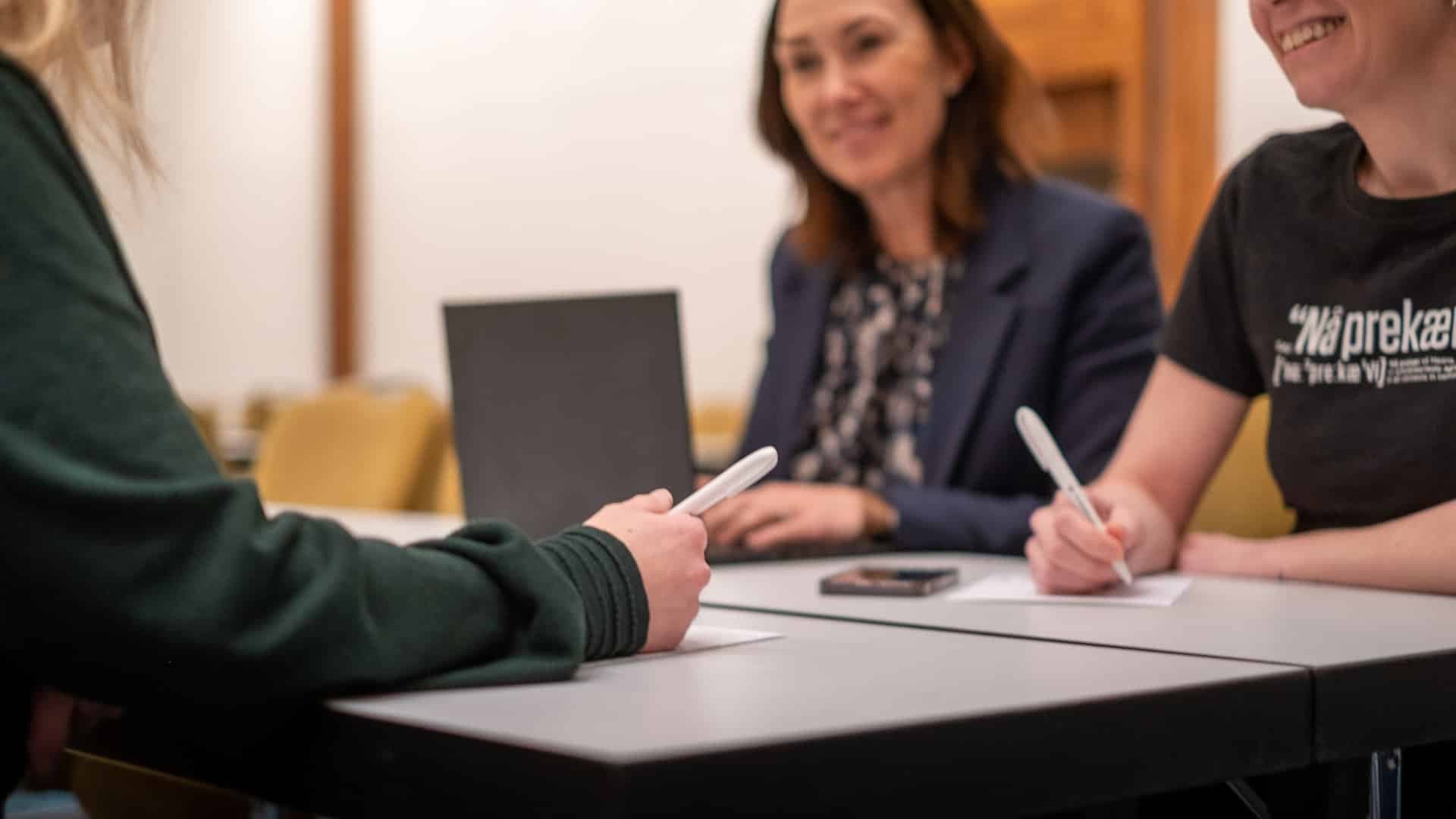 Personer i samtale rundt et møtebord, med notater og PC i et møterom på hotell.